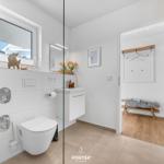Badezimmer mit Toilette, Waschbecken und Dusche hinter Glas. Nebenraum mit Kleiderständer und Bank.