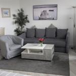 Wohnzimmer mit grauem Sofa, Sitzgelegenheit und Tisch. Wandbild und Pflanze sichtbar.