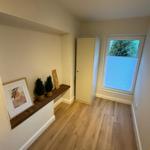 Raum mit Holzboden, Fenster mit Jalousie und kleiner Ablage mit Pflanzen und Bild.