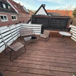 Terrasse mit Holzboden, Sitzmöbeln und Tisch. Umzäunung und Nachbarhäuser im Hintergrund.