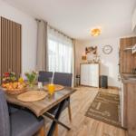 Esszimmer mit Tisch, Stühlen und Küchenbereich. Holzboden und Fenster mit Vorhängen.