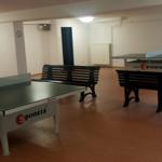 Zwei Tischtennisplatten mit Bänken in einem Innenraum.
