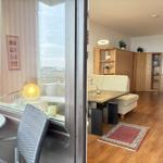 Wohnraum mit Holzboden, Sofa, Tisch und Bücherregal. Durchgang zu Balkon mit Sitzgelegenheit.