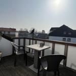 Terrasse mit Tisch, zwei Stühlen und Pflanze. Vogel sitzt auf Pfosten. Blick auf Wohnhäuser.
