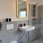 Badezimmer mit Dusche, Waschbecken und Toilette. Graue Fliesen und beleuchtete Spiegel.