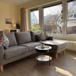 Ein Wohnzimmer mit grauem Sofa, kleinem Tisch und großen Fenstern.