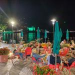 Nachtansicht einer Terrasse mit Gästen, roten Stühlen, grünen Sonnenschirmen und Blick auf einen beleuchteten See.