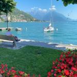 Eine Person sitzt auf einer Bank am Ufer eines Sees mit Booten und Bergen im Hintergrund.