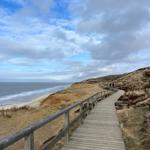 Holzweg führt durch Dünen zur Küste. Blick auf Meer und Strand.