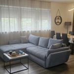 Großer Wohnbereich mit grauem Sofa und Holzboden. Fenster mit weißen Vorhängen.