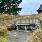 Eine Bank aus Beton und Holz auf einem Steinweg in einer grasbewachsenen Umgebung mit einem Haus im Hintergrund.