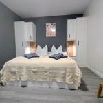 Schlafzimmer mit Bett, Schränken und Holzbank. Wandfarbe grau, Boden hellgrau.