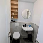 Badezimmer mit Toilette, Waschbecken und Holzregal an der Wand.