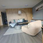 Wohnzimmer mit Sofa, Sessel und Holzwand. Küche und Essbereich sind sichtbar.