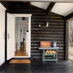 Entrance of a log cabin with open door and stacked firewood. Forest landscape in the background.