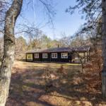 A dark wooden house with white windows stands in the middle of a forest.