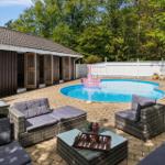 Pool with pink float, patio with seating and wooden house in background.