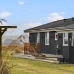 Black wooden house with terrace and garden. Wooden chairs are outside. Sky is blue with white clouds.