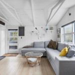 Wohnraum mit grauem Sofa, Glas-Tisch und großen Fenstern. Weiße Wände und Holzboden.