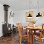Dining room with wooden table, chairs, and wood stove. Wall decor and pendant lights.