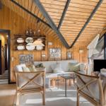 Wohnzimmer mit Holzwand, Sofa, zwei Stühlen und Glas-Tisch. Decke mit Holz und Metallträgern.