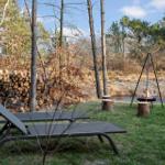 Two lounge chairs next to a grill in the woods. Background: woodpile and river.