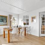 Dining area with wooden table, white chairs, and paintings on walls. Passage to kitchen.