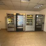 Vier Automaten mit Getränken und Snacks in einem Raum mit Fliesenboden.