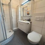 Badezimmer mit Dusche, Waschbecken und Toilette. Fliesenwand und dunkler Boden.