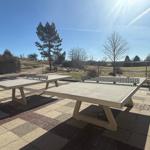 Zwei Tischtennisplatten auf gepflastertem Platz mit Blick auf Bäume und Himmel.