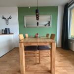 Esszimmer mit Holztisch, Stühlen und grünem Wandbereich. Fenster mit Vorhang und Pflanze.