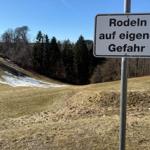 Schild weist auf Schneerodeln hin. Hintergrund: Wald und grasbewachsene Hangflächen.