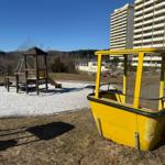 Ein gelbes Fahrgeschäft steht vor einem Spielplatz mit Rutsche und Klettergerüst.