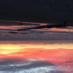 Sonnenuntergang am Strand mit Reflexion im Wasser und Silhouetten von Personen.