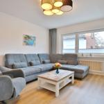 Wohnzimmer mit grauem Sofa, Sessel und Holztisch. Fenster mit Jalousien und Wandbild.