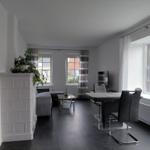 Wohn- und Essbereich mit Sofa, Tisch und Stühlen. Große Fenster mit Blick nach draußen.