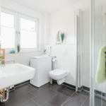 Badezimmer mit Dusche, Waschbecken und Toilette. Fenster, graue Fliesen und weiße Wände.
