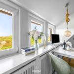 Schreibtisch mit Sicht auf Dünenlandschaft, Blumen, Bücher und Lampe.