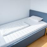 Ein Bett mit weißer Matratze und Kissen. Neben dem Bett steht ein kleiner Tisch mit einer Lampe.