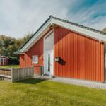 Rotes Holzhaus mit weißer Tür und Garten. Spielplatz und kleiner Schuppen im Hintergrund.