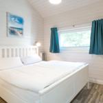 Schlafzimmer mit weißem Bett, Holzwand und Fenster mit blauen Vorhängen.