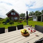Holztisch mit Obst und Wein auf Terrasse vor rotem Haus mit Spielplatz.