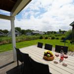 Terrasse mit Tisch, Stühlen, Obst und Wein. Blick auf grünen Garten und roten Haus.