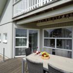 Terrasse mit Tisch, Stühlen und Obst. Hintergrund: Haus mit Fenstern und Balkon.