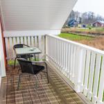 Terrasse mit Tisch und zwei Stühlen. Weißes Geländer und Holzboden. Blick auf Grünland und Häuser.