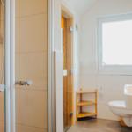 Bad mit Dusche, Waschbecken und Toilette. Holzregal und Fenster sichtbar.