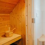 Holzsauna mit Sitzbank und Eimer. Türöffnung zeigt weiße Wand.