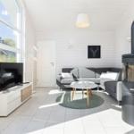 Wohnzimmer mit grauem Sofa, Holztisch und Kaminofen. Große Fenster und weißer Holzboden.