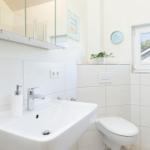 Badezimmer mit Waschbecken, Toilette und Fenster. Spiegel, Pflanze und Uhr sind sichtbar.