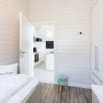 Zimmer mit Bett, Tür zum Küchenbereich und Stuhl. Holzwände und weißer Schrank.